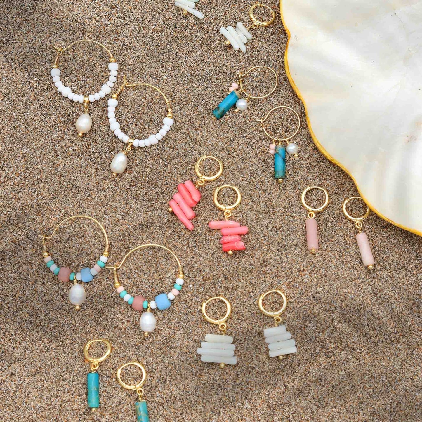 Assorted beach-style earrings including GIADA PECAHAN WIRE HOOP EARRINGS on sand, showcasing colorful beads and pearl accents.