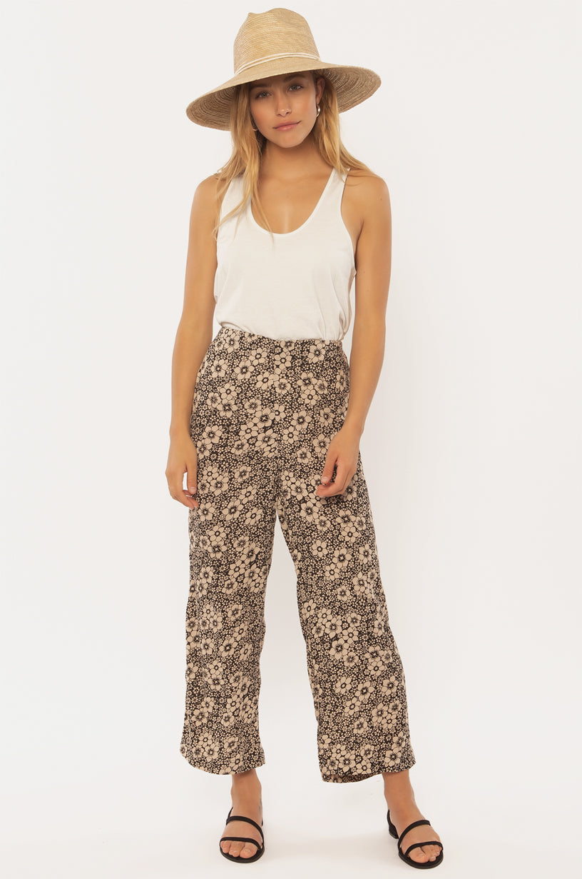 Sisstrevolution Dusty Skies WVN Pant in black floral print, paired with a white tank top and straw hat.