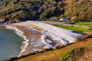 Dan Santilo Glass Placemat - Pwll Du features a stunning coastal photo of Pwll Du beach with white surf, green hills, and seaside cottages. Made from toughened glass, this 22.5x18.5cm placemat adds scenic charm to any dining table.