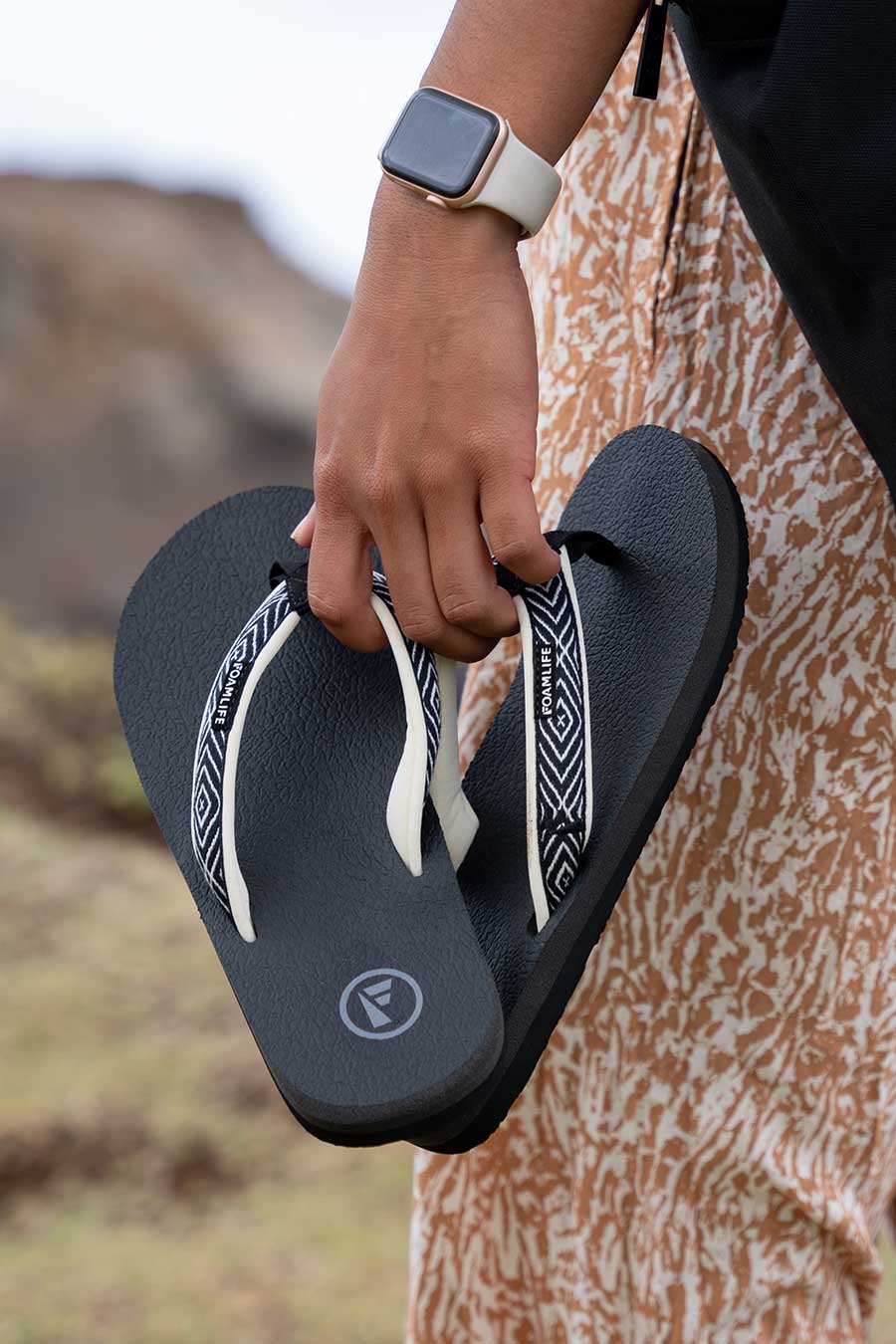 Hand holding FoamLife Yogi black flip flops with stylish patterned straps.