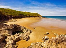 Scenic beach view with golden sand, rocky foreground, and grassy hills under a blue sky.