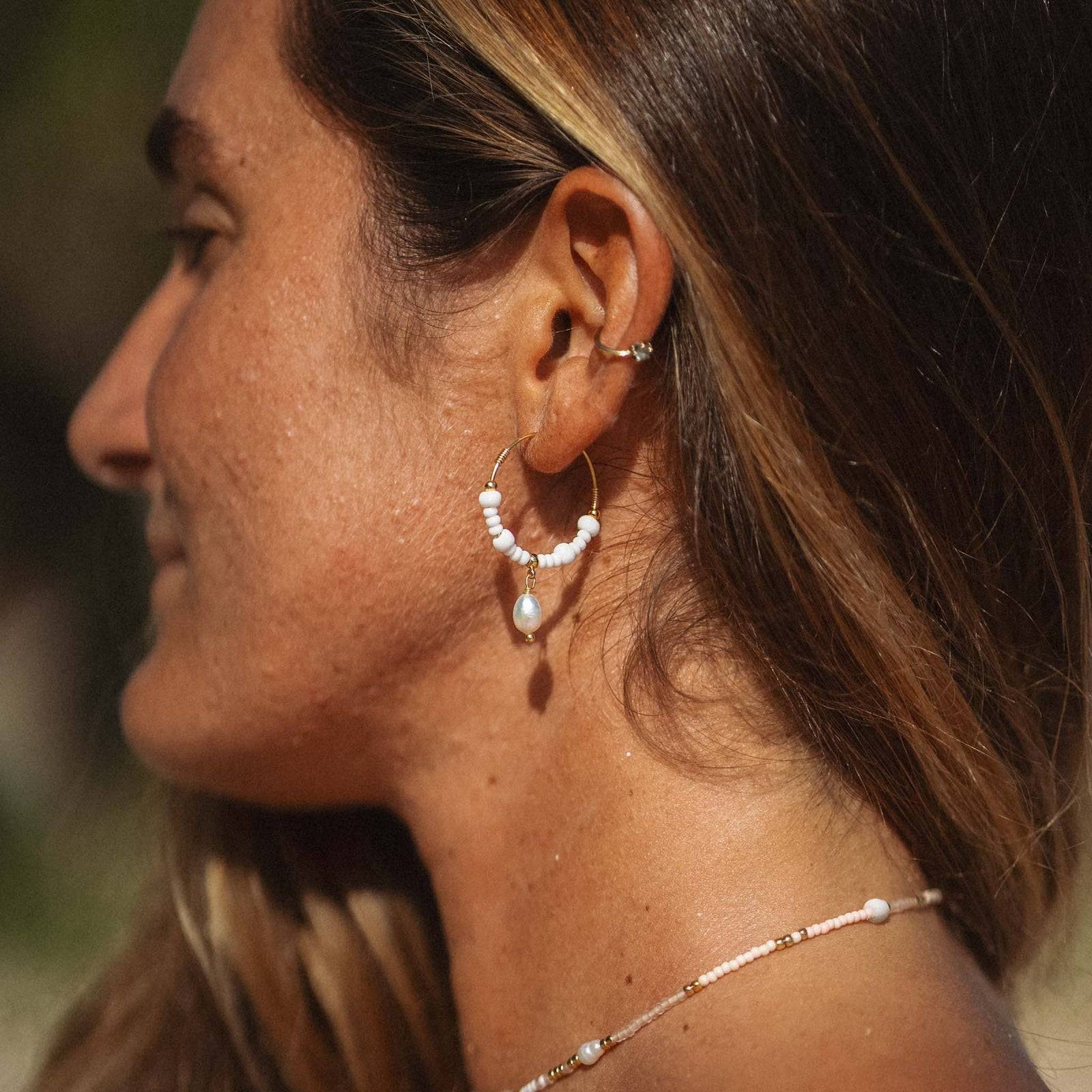GIADA GEGAMBAR WIRE HOOP EARRINGS styled on model outdoors, highlighting the white beads and pearl drop in a beach setting.