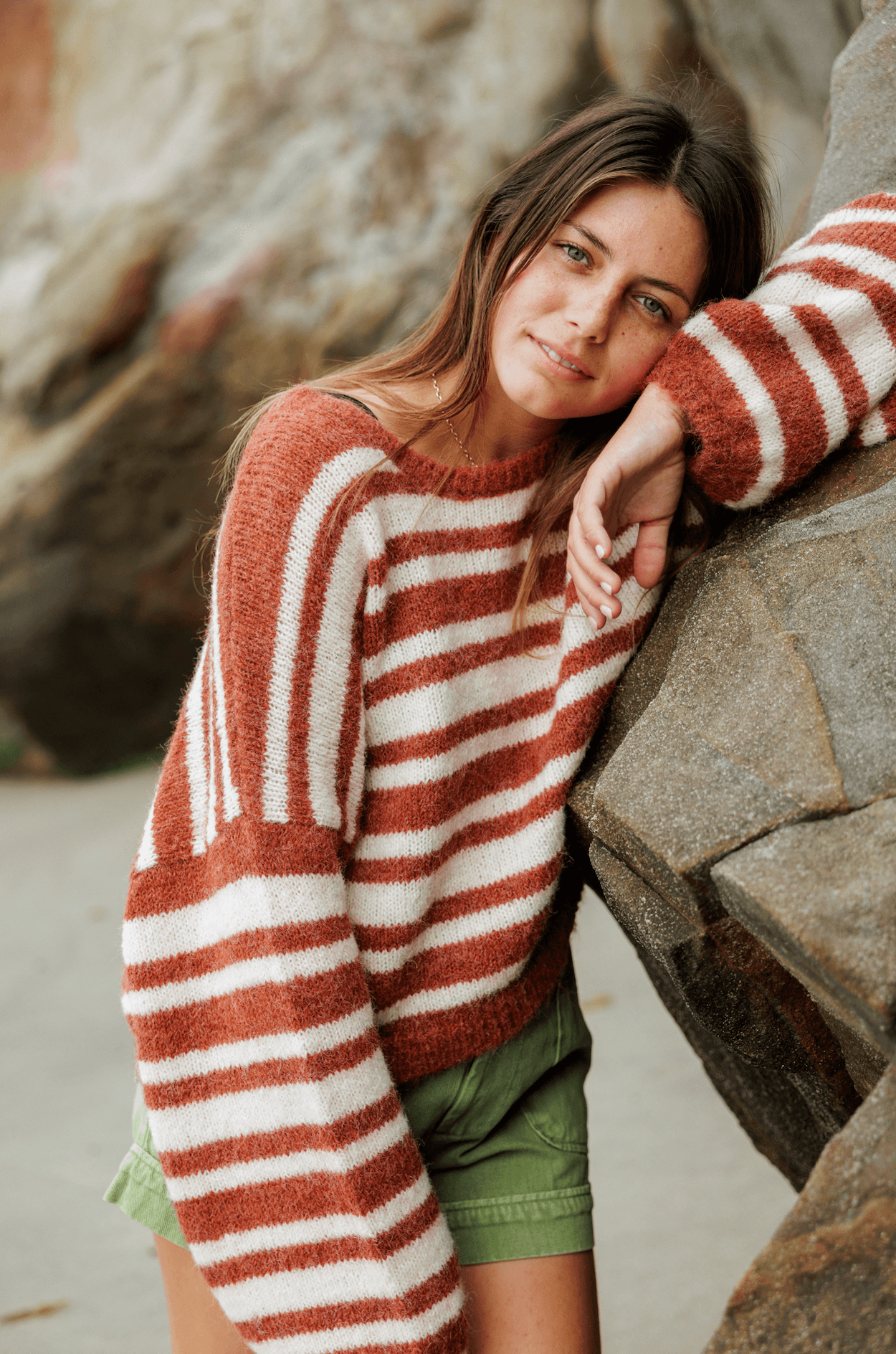 Cozy Sisstr Ocean Shores Sweater in rust, paired with green shorts near rocks.