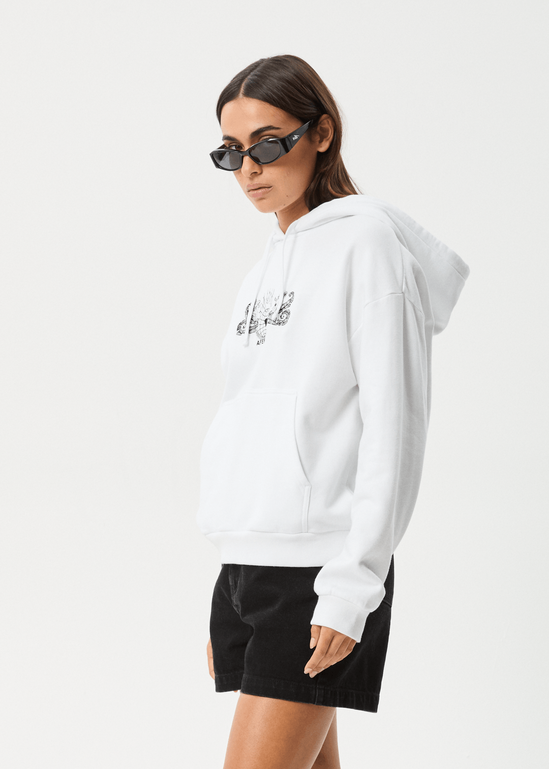 Model wearing black rectangular sunglasses and a white hoodie with a front pocket and graphic print, paired with black shorts, against a plain background.
