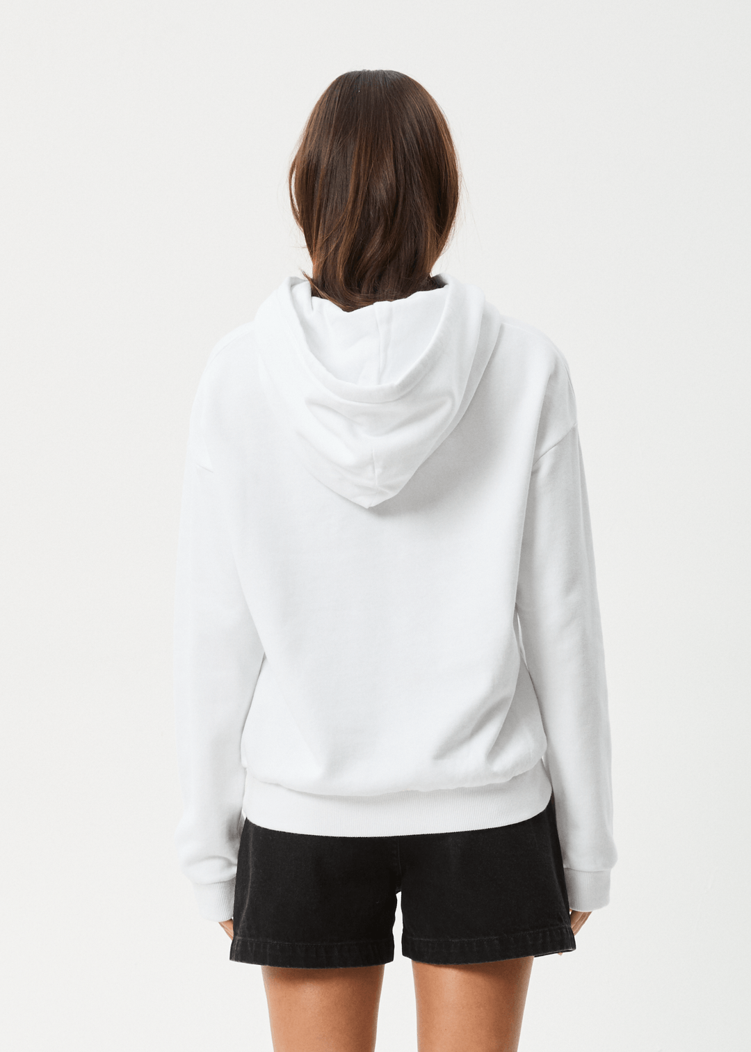 Back view of a woman wearing a white hoodie with a relaxed fit, drawstring hood, front pouch pocket, and black shorts, plain background