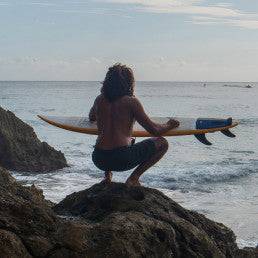 Surfer holding a surfboard with Deflow Adrien Toyon pad, overlooking the ocean.