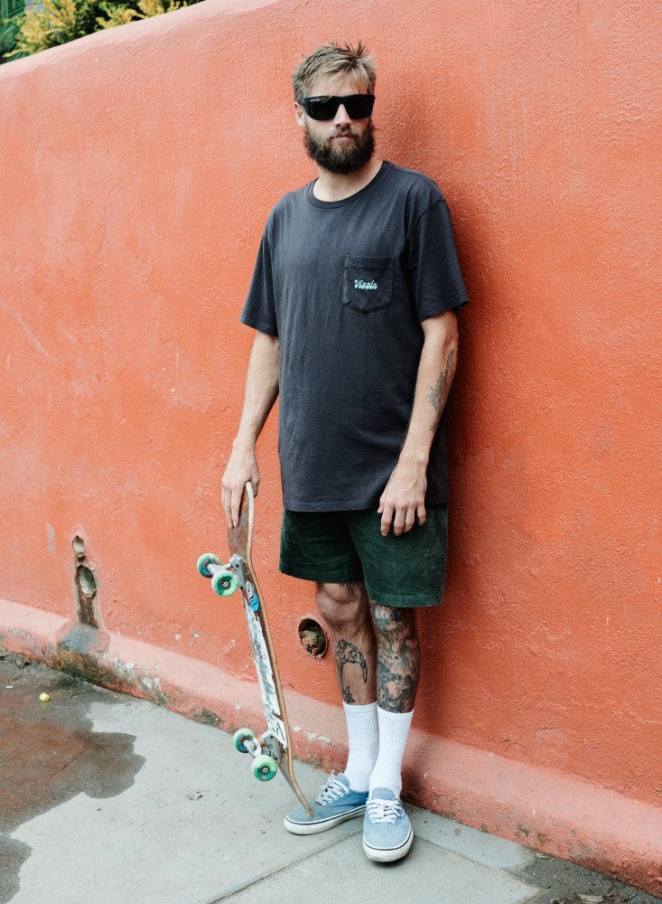 Man wearing I-sea Greyson 2.0 black sunglasses holding a skateboard near an orange wall.