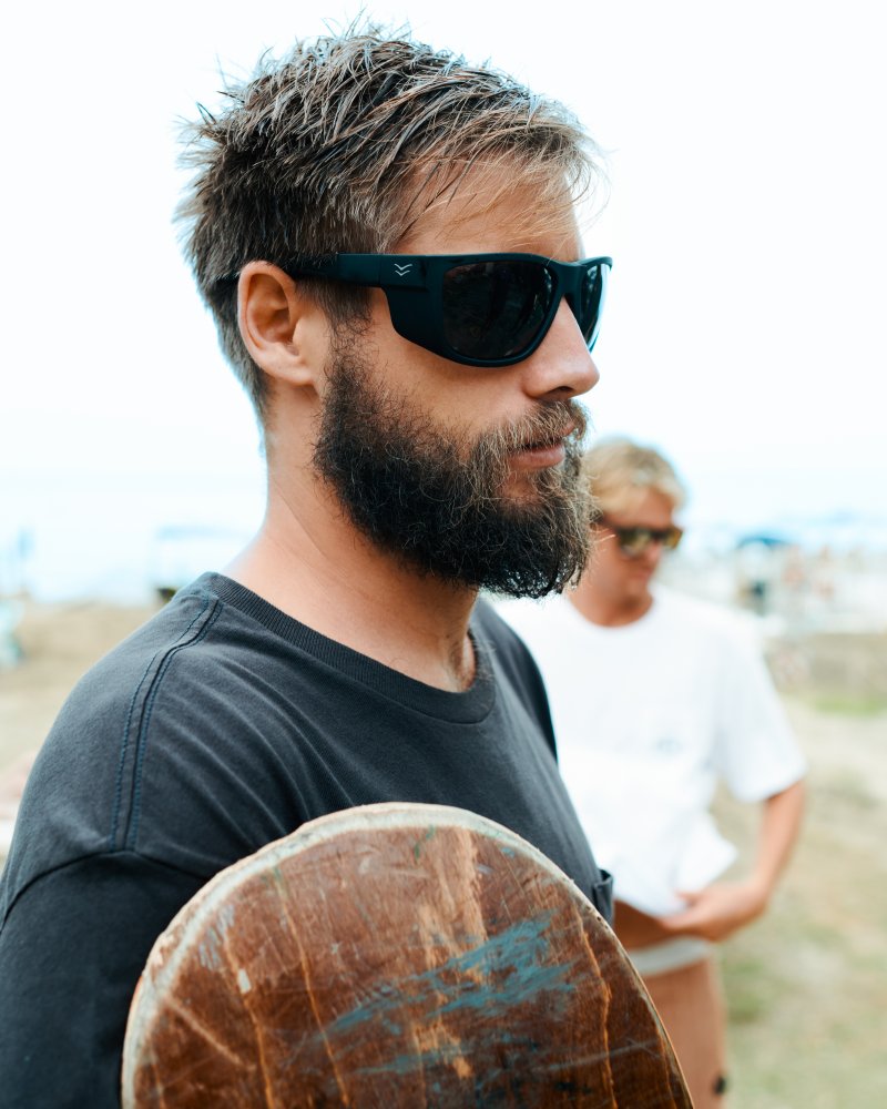 Man wearing I-sea Greyson 2.0 black sunglasses holding a skateboard outdoors.