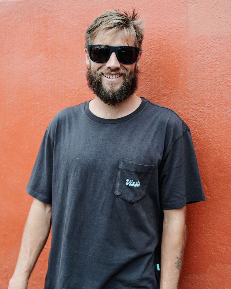 Man smiling and wearing I-sea Greyson 2.0 black sunglasses against an orange wall.